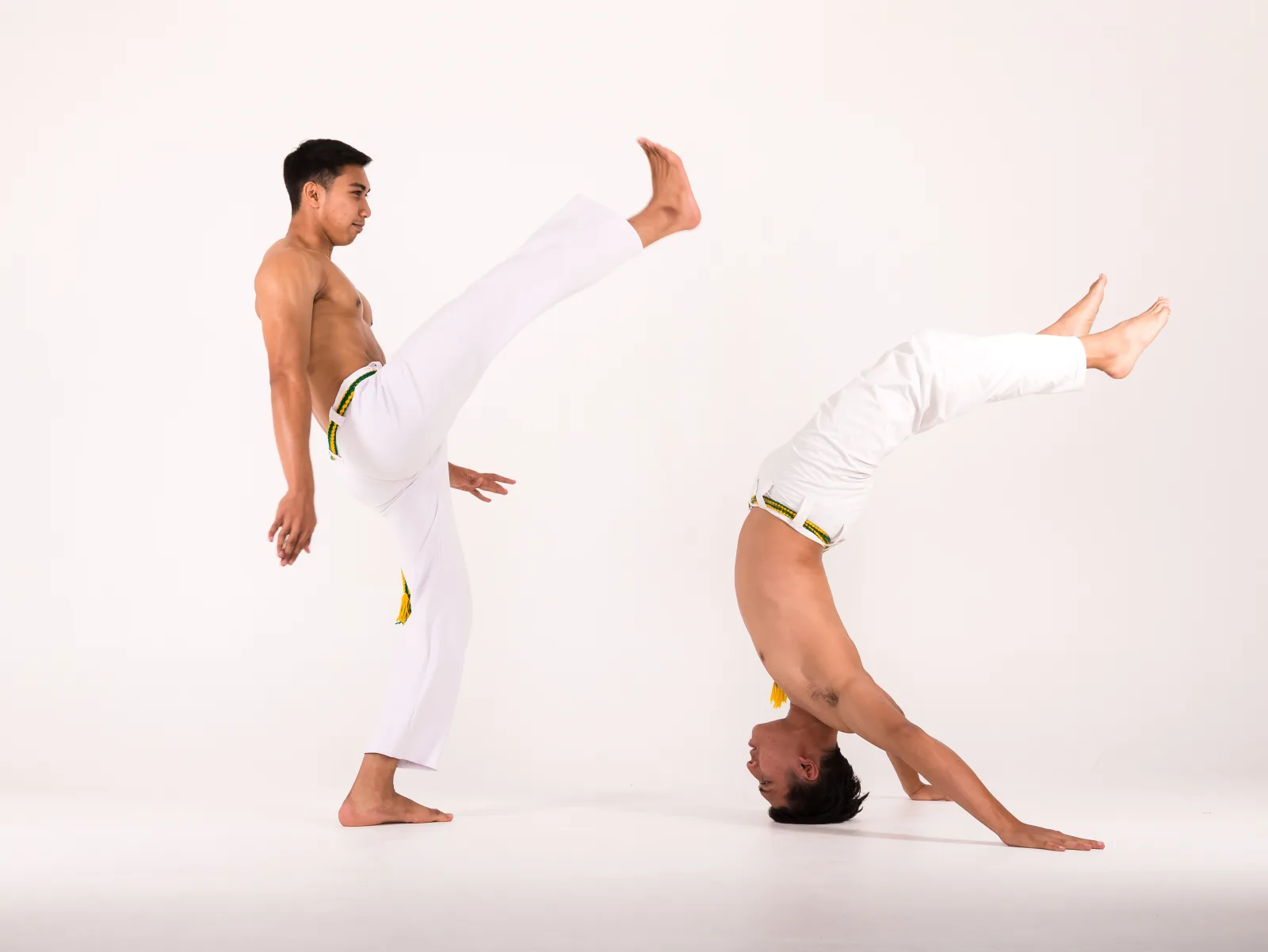 Two Capoeira practitioners in white trousers demonstrate a high kick and an inverted balance in a studio.