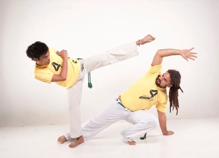 Two Capoeira practitioners demonstrate a kick and an evasive floor movement in a studio.