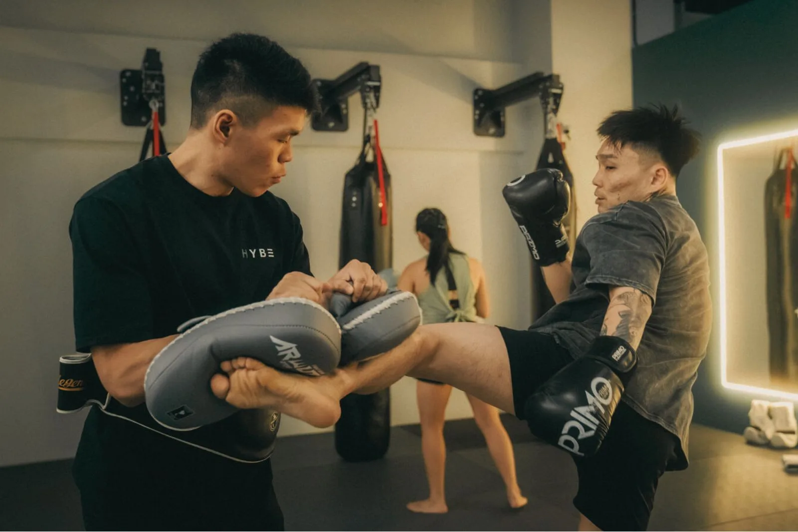 A HYBE coach holds pads while a Muay Thai student lands a kick.