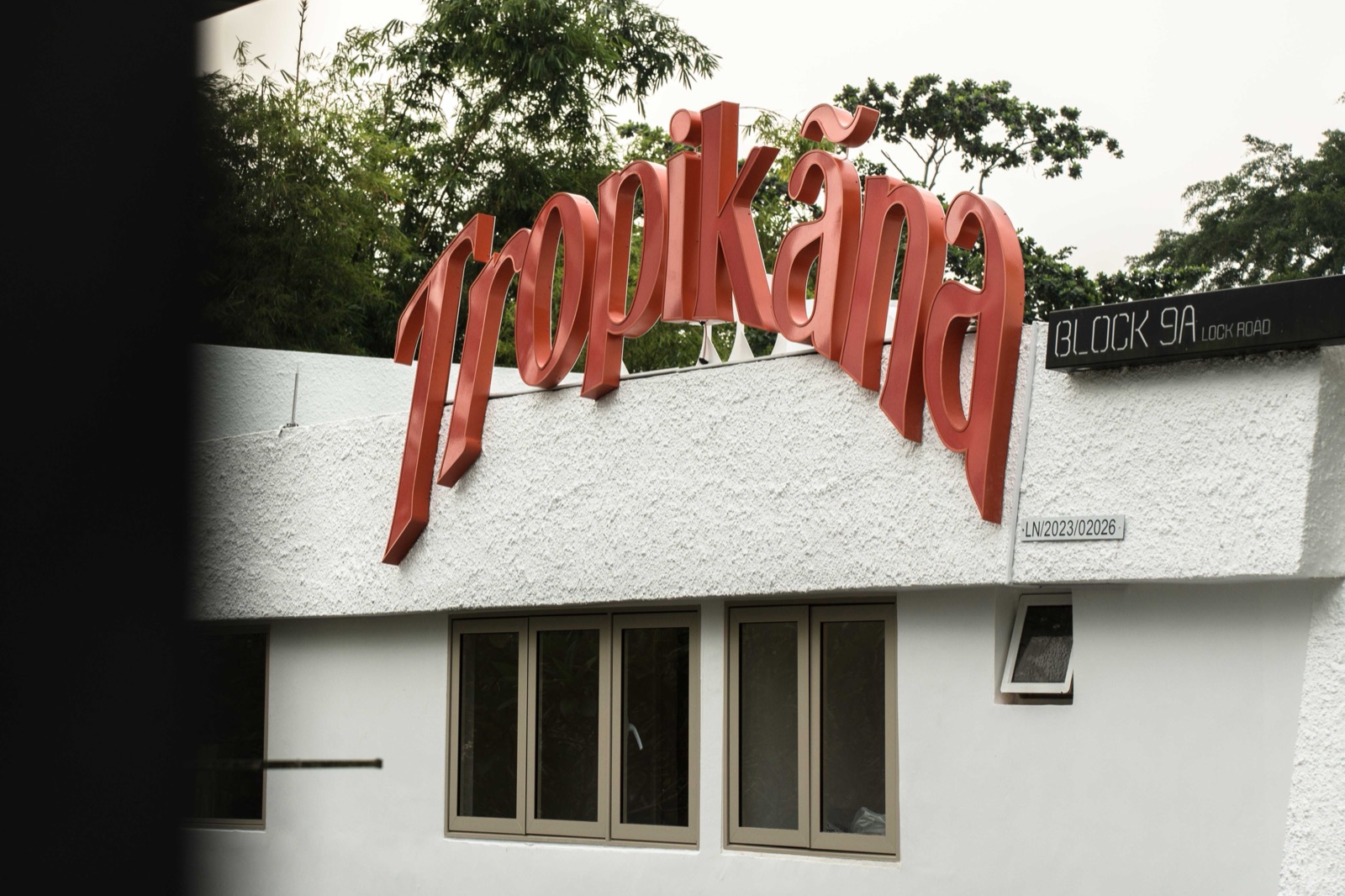 The Tropikãna sign in red script standing on the white wall of the building, with a Block 9A Lock Road street sign beside it